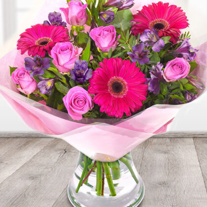 The Best Flowers for International Women's Day is a delightful bouquet featuring a blend of vibrant pink Gerbera Daisies, pink Roses, and elegant white Lily buds, with soft greenery and small white filler flowers. Arranged in a hand-tied bouquet wrapped in pink paper with a pink mesh ribbon, it’s the perfect gift for general occasions, celebrations, or as a gesture of appreciation or congratulations