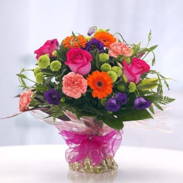Gerbera Daisy and Rose Arrangement with soft Carnations 1 Rose and Gerbera Daisy Bouquet featuring bright pink roses, striking orange Gerbera daisies, peach carnations, and deep purple accent flowers, beautifully wrapped with a sheer pink ribbon.