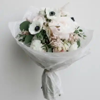 Hand-tied anemone and peony bouquet wrapped in white textured paper with fresh greenery and rustic twine.