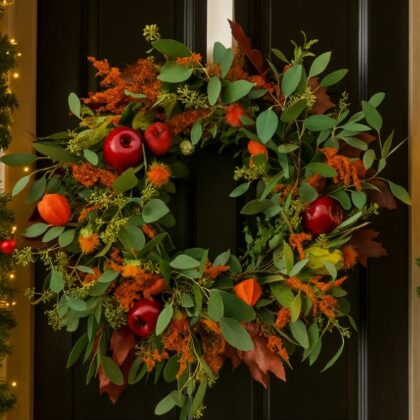 A close-up shot of a rustic, handmade autumn door wreath featuring faux orange, red, and yellow leaves and small berries.