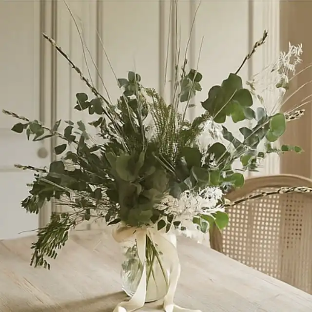 Blog 2 A lush, airy bouquet of assorted green and dried white foliage, eucalyptus, and tall grasses in a glass vase tied with a ribbon. Christmas foliage bouquet for sales