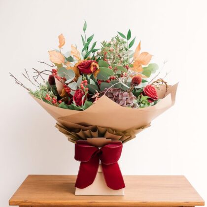 Rustic-style bouquet with deep red roses, eucalyptus, autumn leaves, red berries, and foliage, wrapped in brown paper and presented in a box with a velvet red bow. Send Christmas Flowers Bouquet Next day delivery