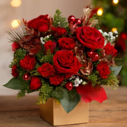 Festive flowers for Christmas delivery featuring red roses, carnations, red baubles, and pine sprigs in a golden decorative box, with a blurry Christmas tree background. Luxury Christmas Hat Box Flowers for sale