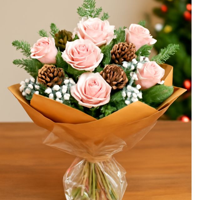 Hand-tied pink Christmas bouquet with fresh pink roses, snowy accents, natural pine cones, and pine foliage, wrapped in a brown paper cone.