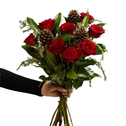 A hand holding a vibrant bouquet of deep red roses mixed with pine sprigs and natural pine cones, perfect for holiday flower arrangements Christmas. Red Rose Christmas Bouquet for sale