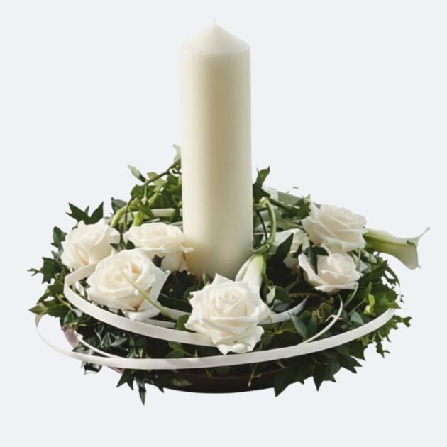 White Rose and Pillar Candle Wreath Centrepiece 1 Candle Flower Wreath featuring a tall white pillar candle, encircled by a wreath of fresh white roses, lush ivy greenery, and delicate white ribbons, perfect for a wedding or memorial table setting.